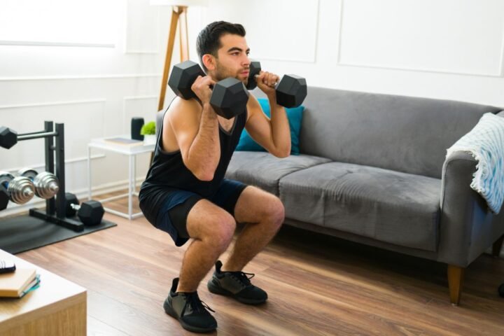 Man performs squat in his living room, holding dumbbells by his shoulders