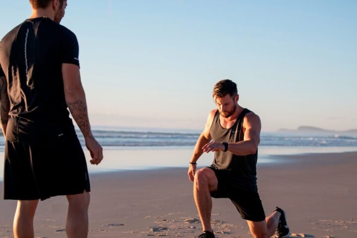 Chris Hemsworth performing lunge exercises on the beach, his trainer Luke Zocchi looks on