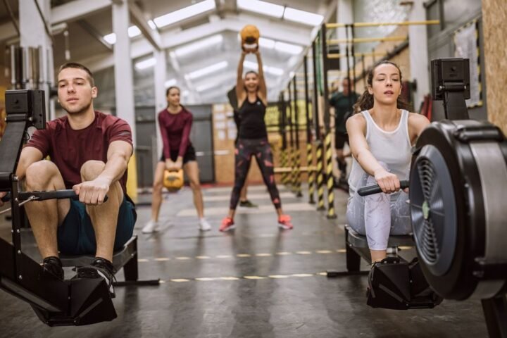 Two people rowing on rowing machines in the foreground, two people working out with kettlebells in the background